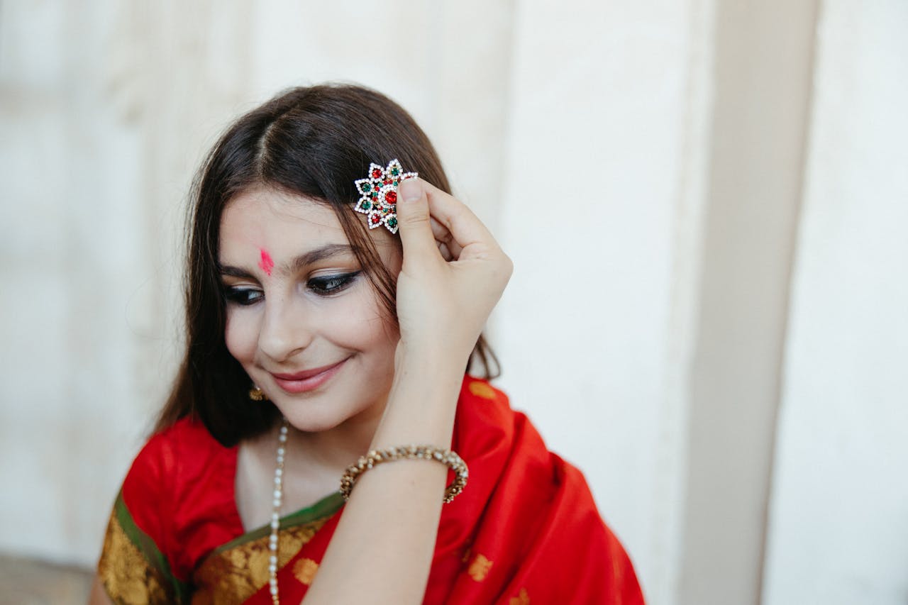 Woman in a red sari with bindi, wearing a traditional hair accessory with a delicate hand adjusting it.